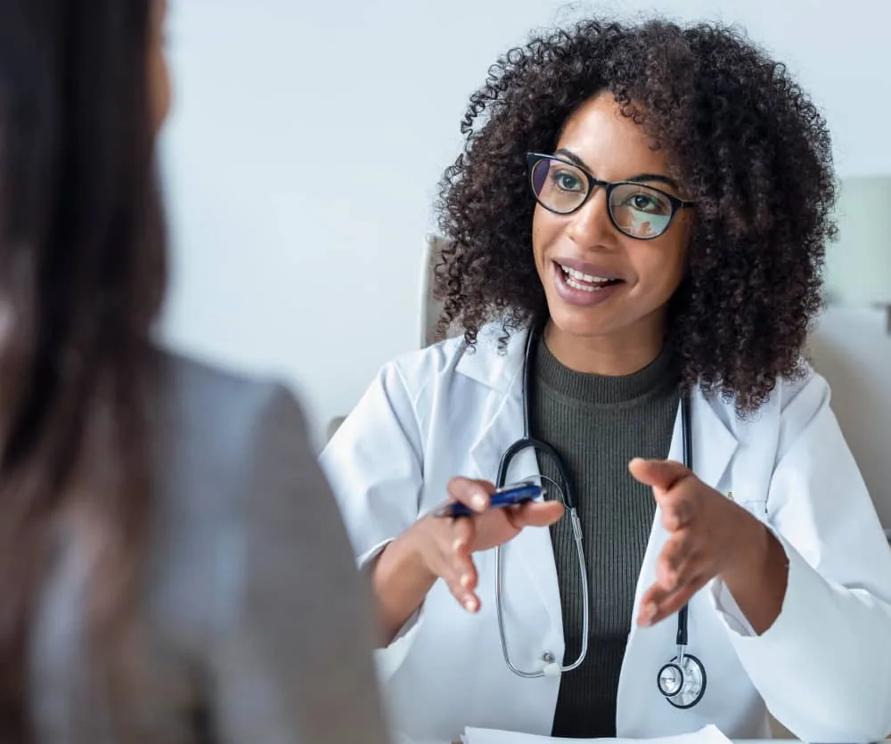 A smiling female doctor wearing a white lab coat, glasses, and a stethoscope around her neck, holding a pen and gesturing with both hands while speaking to a patient during a medical consultation.
