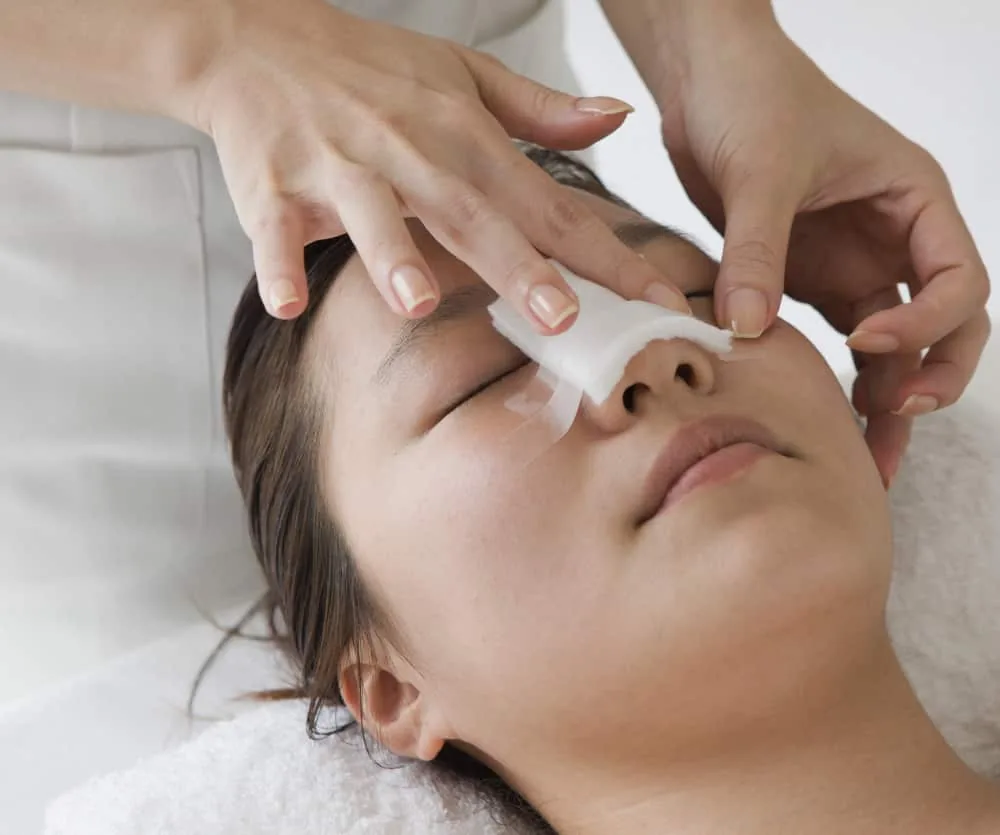 A woman lying on a treatment table with eyes closed while a practitioner applies a white post-surgical or medical dressing over the nose and eye area, commonly used after rhinoplasty surgery to manage swelling.