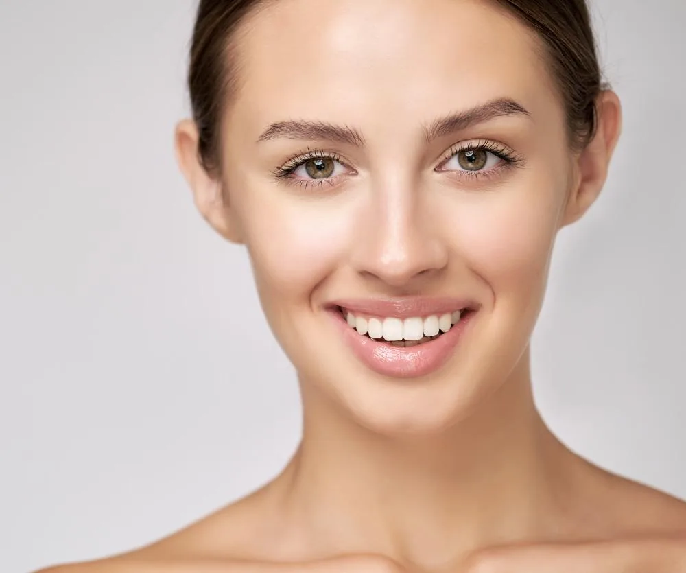 A young woman with clear, smooth skin, hazel eyes, and dark hair pulled back, smiling naturally with white teeth, showcasing a well-defined jawline and neck — illustrating a neck lift recovery result.