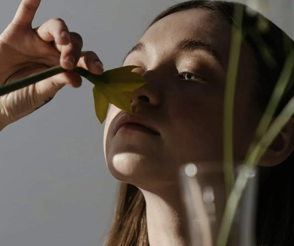 A young woman with dark hair holding a yellow flower stem close to her nose with her eyes gently closed, with green plant stems visible in the foreground, in a soft natural light setting — illustrating a rhinoplasty surgery preparation article.