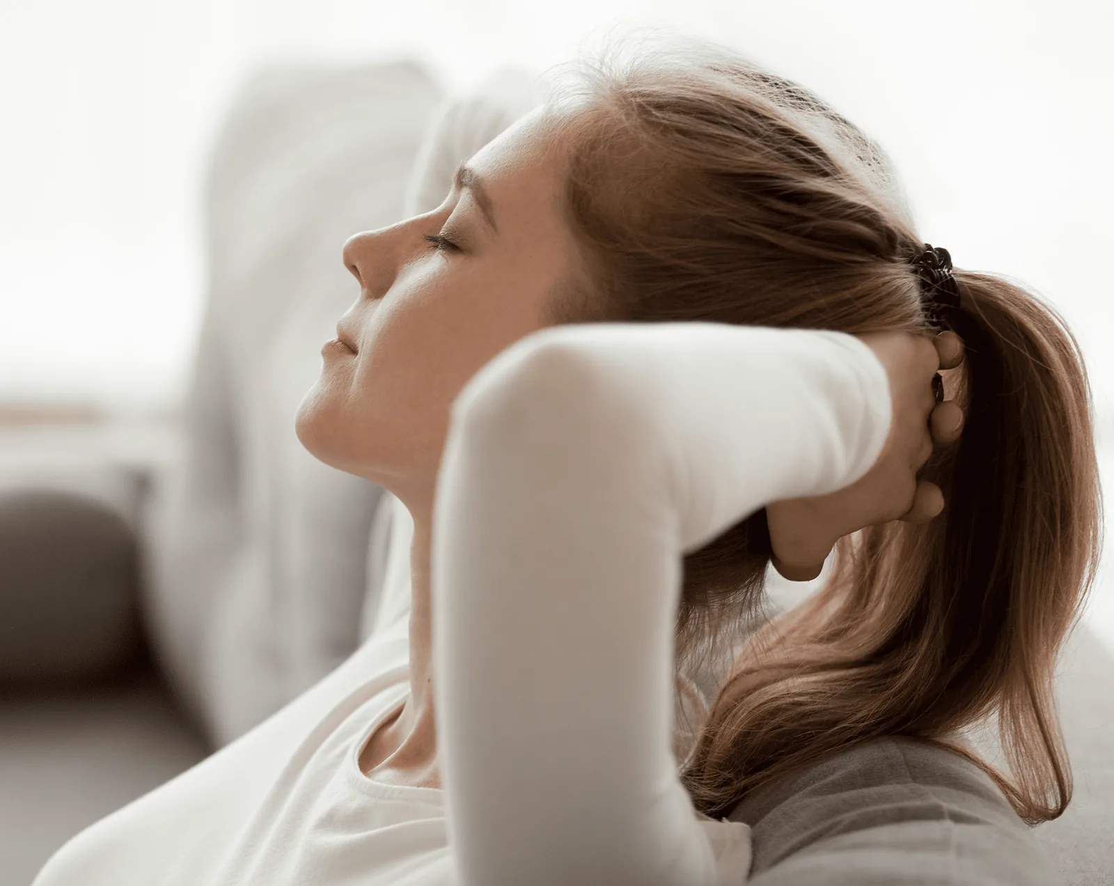 A young woman with brown hair in a ponytail, wearing a white top, leaning back on a sofa with her eyes closed and hands clasped behind her head in a relaxed, peaceful pose.