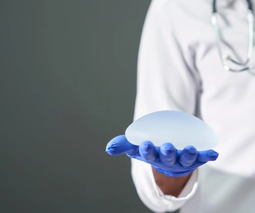 A medical professional in a white lab coat and blue latex gloves holds a silicone breast implant in the palm of their hand, with a stethoscope visible around their neck.