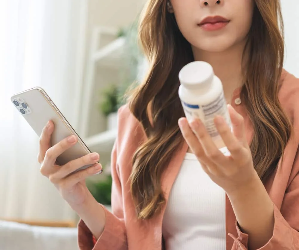 A young woman with long brown hair wearing a pink cardigan over a white top, holding a smartphone in one hand and a white supplement or medication bottle in the other, appearing to research the product's information.