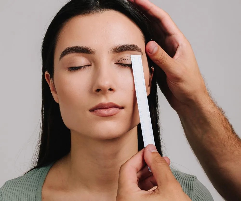 A woman with closed eyes and dark hair wearing a sage green top, with surgical dotted markings drawn around her right eyelid area, while a practitioner holds a white measurement strip vertically against her eye and uses the other hand to gently lift her
