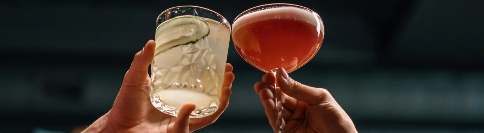 Two hands clinking cocktail glasses in a toast — one hand holding a crystal rocks glass with a clear drink garnished with a cucumber slice, and the other holding a coupe glass filled with a pink cocktail.