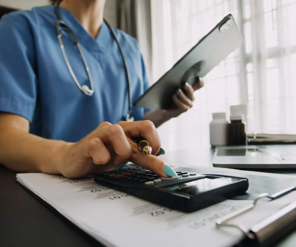 A healthcare professional in blue scrubs with a stethoscope around their neck using a calculator while holding a clipboard, with medical documents on the desk and medication bottles in the background, representing the cost of labiaplasty surgery.