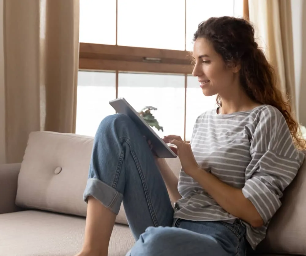 A young woman with curly auburn hair smiling while using a tablet, relaxing on a beige sofa in a bright living room, wearing a gray striped top and rolled-up blue jeans, with a window and curtains in the background.