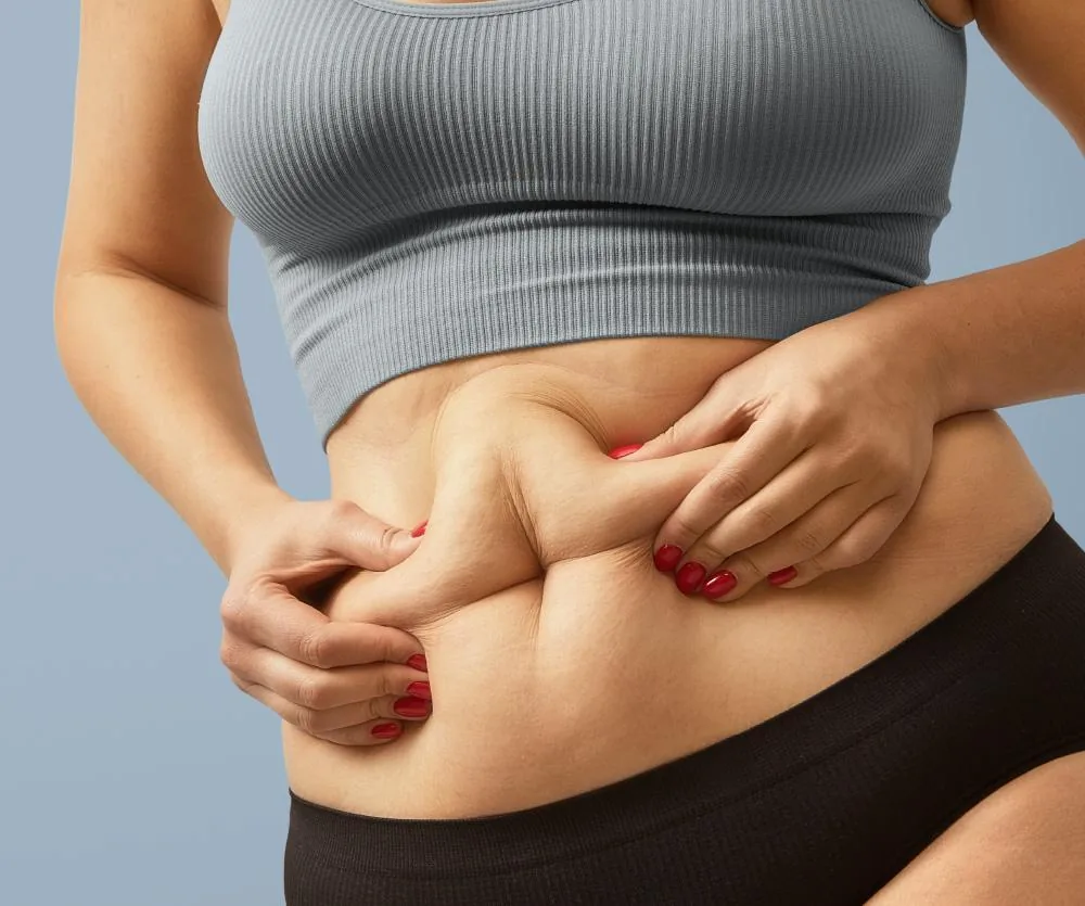A woman in a gray ribbed crop top and black shorts pinching excess abdominal skin and belly fat with both hands, illustrating loose skin or body fat on the midsection, relevant to topics such as skin removal surgery or body contouring.