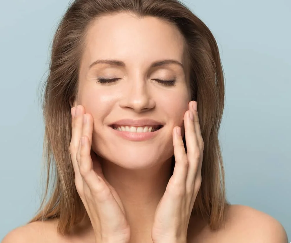 A smiling woman with closed eyes and long brown hair gently touching both sides of her face with her fingertips, showcasing smooth, healthy skin — commonly used to illustrate buccal fat removal results.