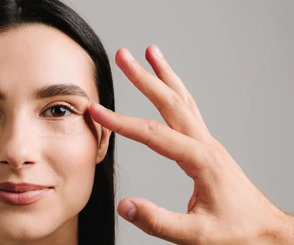 A woman with dark hair gently touching the outer corner of her eye with one finger, highlighting the eye area, relevant to a discussion about how long eyes feel tight after blepharoplasty (eyelid surgery).
