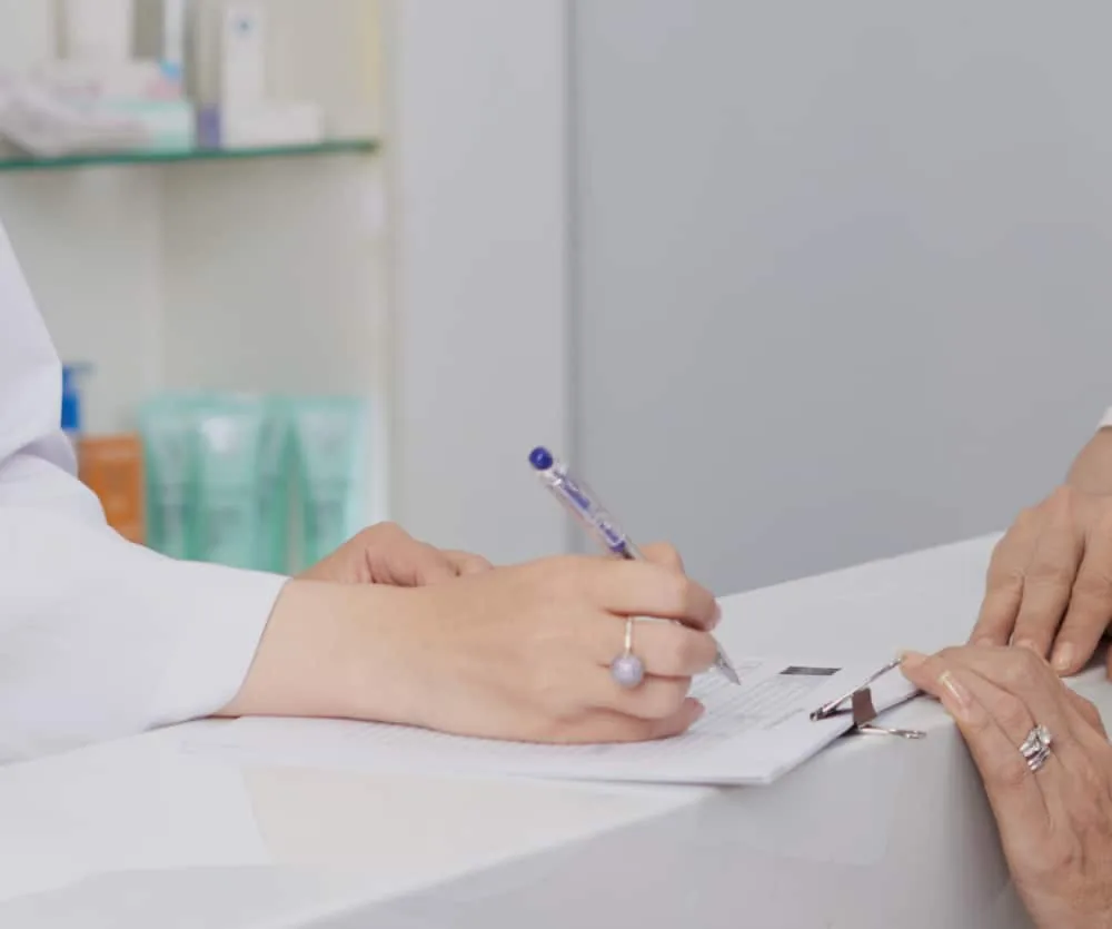 A healthcare professional in a white coat writing on a clipboard with a blue pen during a consultation, while a patient's hands rest on the desk across from them. Medical and skincare products are visible on shelves in the background.
