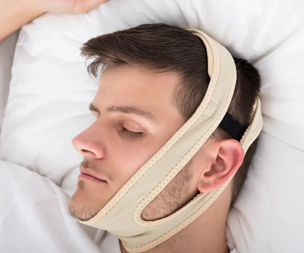 A man sleeping in bed on a white pillow wearing a beige chin strap support wrap around his jaw and head, commonly used after a neck lift procedure or to reduce snoring.