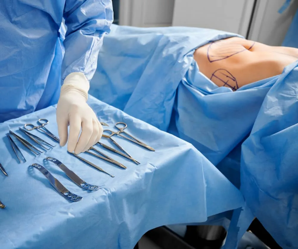 A surgeon in a blue gown and white latex gloves reaches for sterile surgical instruments laid out on a blue drape, while a patient's abdomen is visible with pre-operative marker lines drawn on the skin, indicating preparation for a mummy makeover plastic