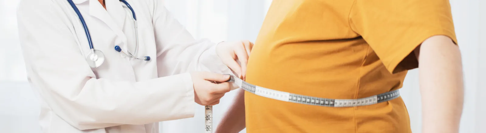 A doctor in a white coat with a stethoscope measuring the waist of a patient wearing a mustard yellow short-sleeve shirt using a tape measure, illustrating a body measurement consultation related to weight loss after liposuction.