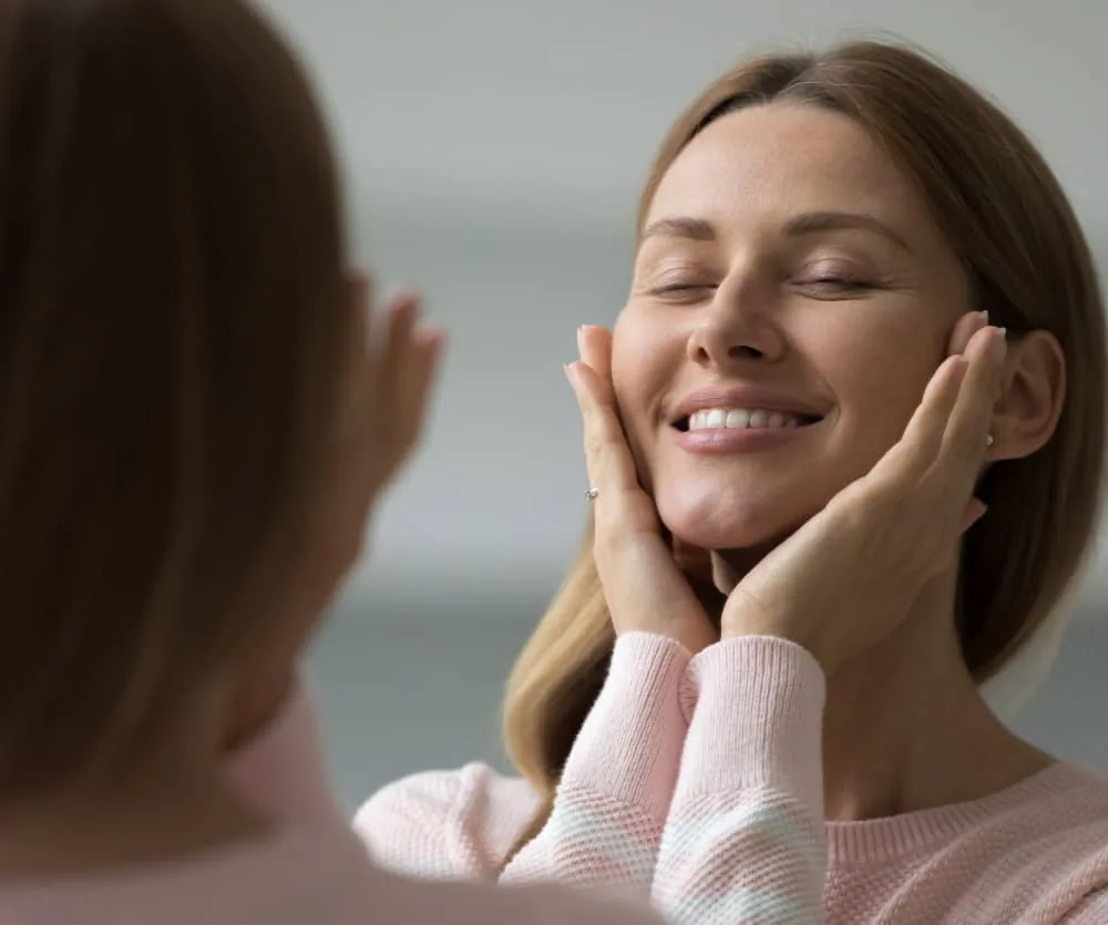 A smiling young woman with closed eyes gently touching her smooth face with both hands, wearing a pink sweater, seen reflected in a mirror — illustrating skin care preparation for laser hair removal.