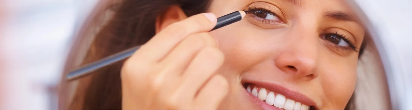 A smiling woman applying eyeliner pencil near her eye, illustrating tips for safely applying eye makeup, relevant to post-blepharoplasty care.