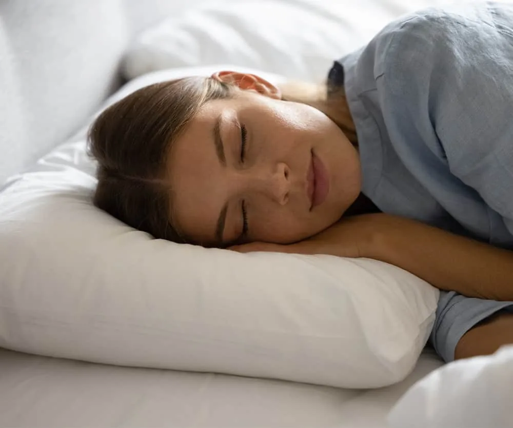 A young woman with brown hair sleeping peacefully on a white pillow, wearing a light blue shirt, lying on white bedding with her eyes closed and arms folded beneath her head.