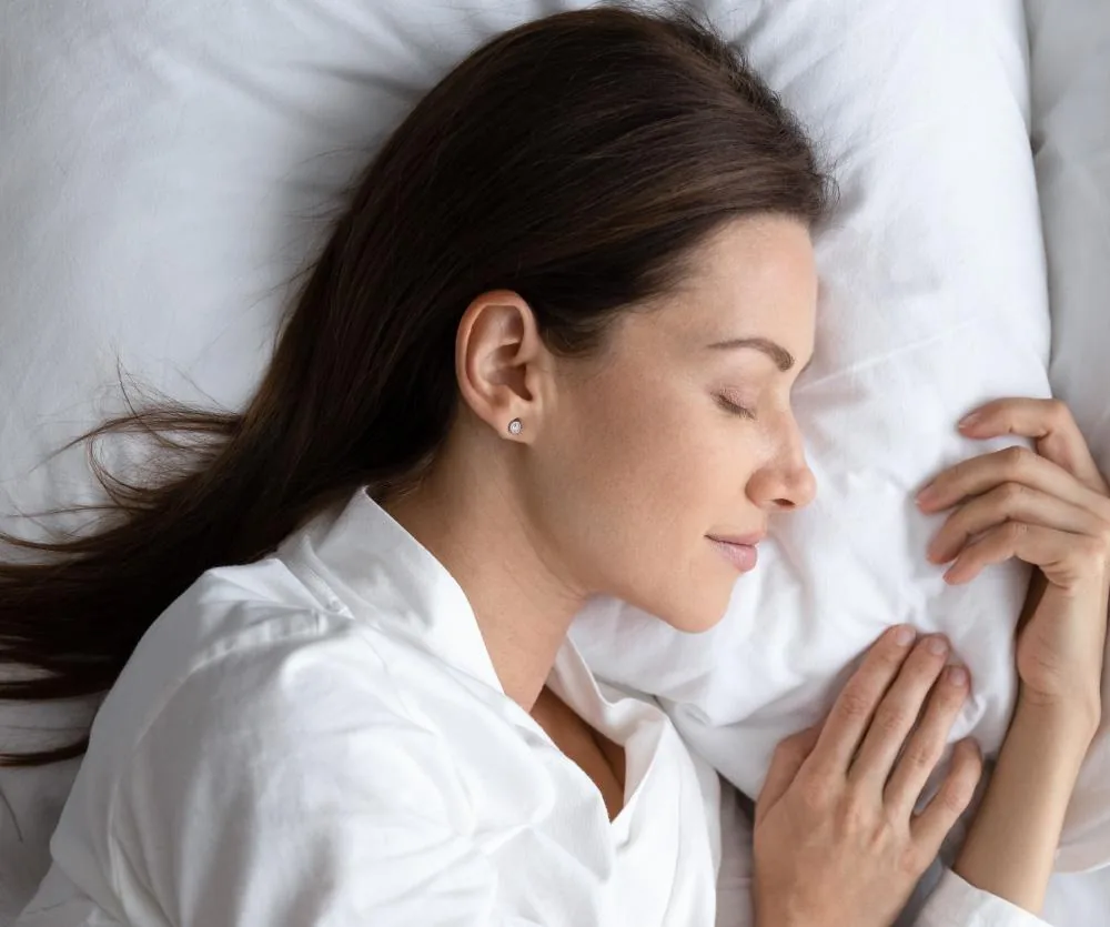 A woman with long dark hair sleeping peacefully on her side on a white pillow, wearing a white shirt and small stud earrings, with her hands gently resting near her face.