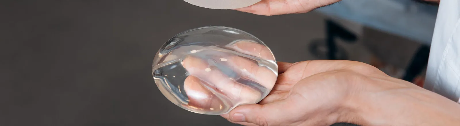 A person holding a clear silicone gel breast implant in both hands, with a second implant partially visible at the top of the frame, illustrating breast augmentation surgery implants.