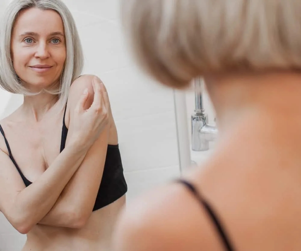 A middle-aged woman with short silver-blonde hair and blue eyes wearing a black bralette, standing in a bathroom and looking at her reflection in a mirror while touching her shoulder, appearing confident and at ease.