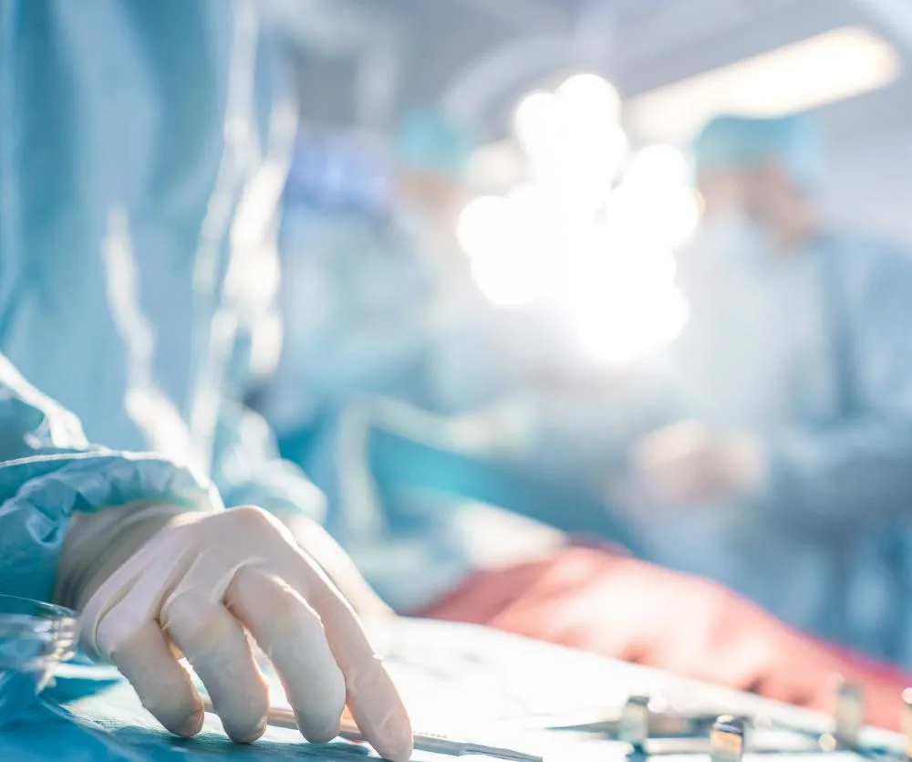 A gloved surgeon's hand resting on a surgical drape in an operating room, with blurred medical team members and bright overhead surgical lights visible in the background, representing the risks associated with a Brazilian Butt Lift procedure.