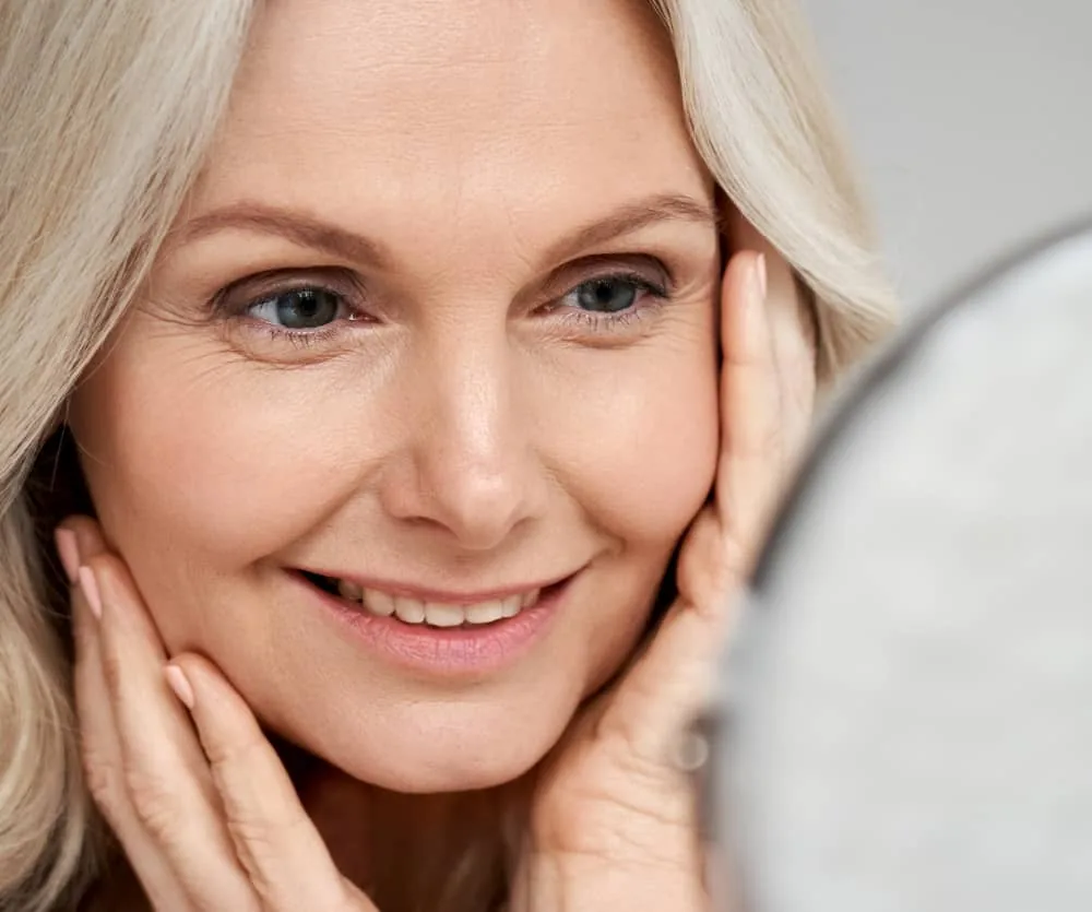 A middle-aged woman with blonde hair and blue eyes smiling warmly while gently touching both sides of her face with her hands, examining her skin in a handheld mirror — related to buccal fat removal and facial aging.