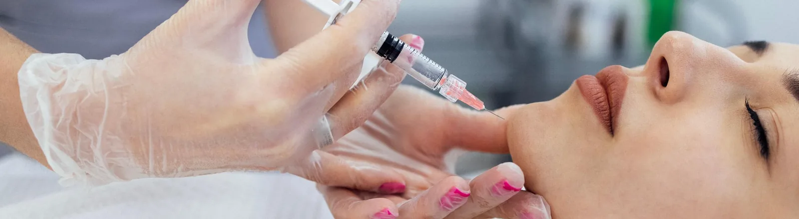 A close-up of a woman with eyes closed receiving a Botox or dermal filler injection to the side of her face, administered by a gloved medical professional holding a syringe.
