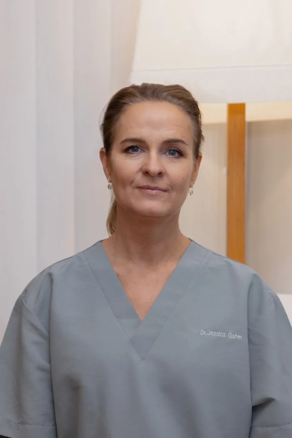 Dr. Jessica Gahm, a woman with blonde hair pulled back, wearing gray medical scrubs embroidered with her name, photographed in a warmly lit clinical interior.