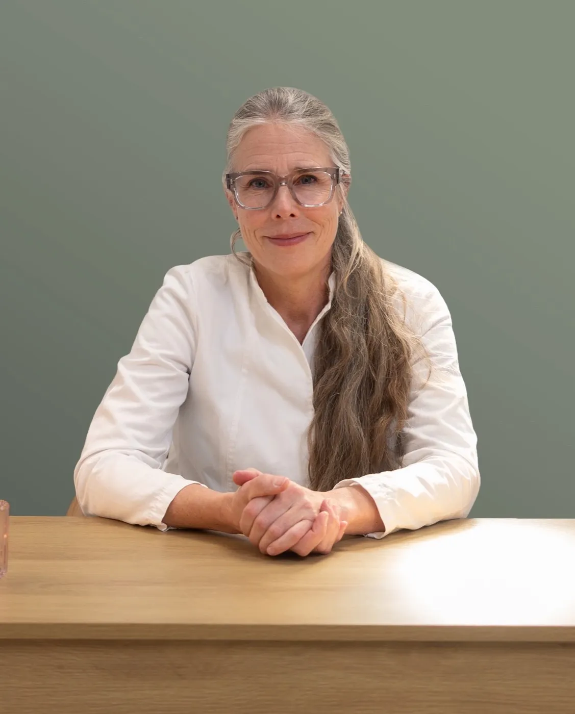 Dr. Stina Klasson, a woman with long gray hair and glasses, wearing a white medical shirt, seated at a wooden table against a muted green background.