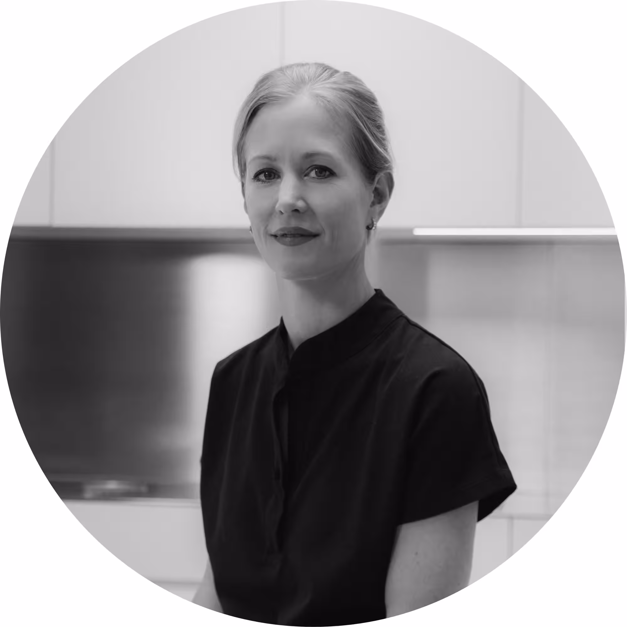 Black and white circular portrait photo of a woman with her hair pulled back, wearing a black short-sleeve top, looking calmly at the camera, with a modern interior setting in the background.