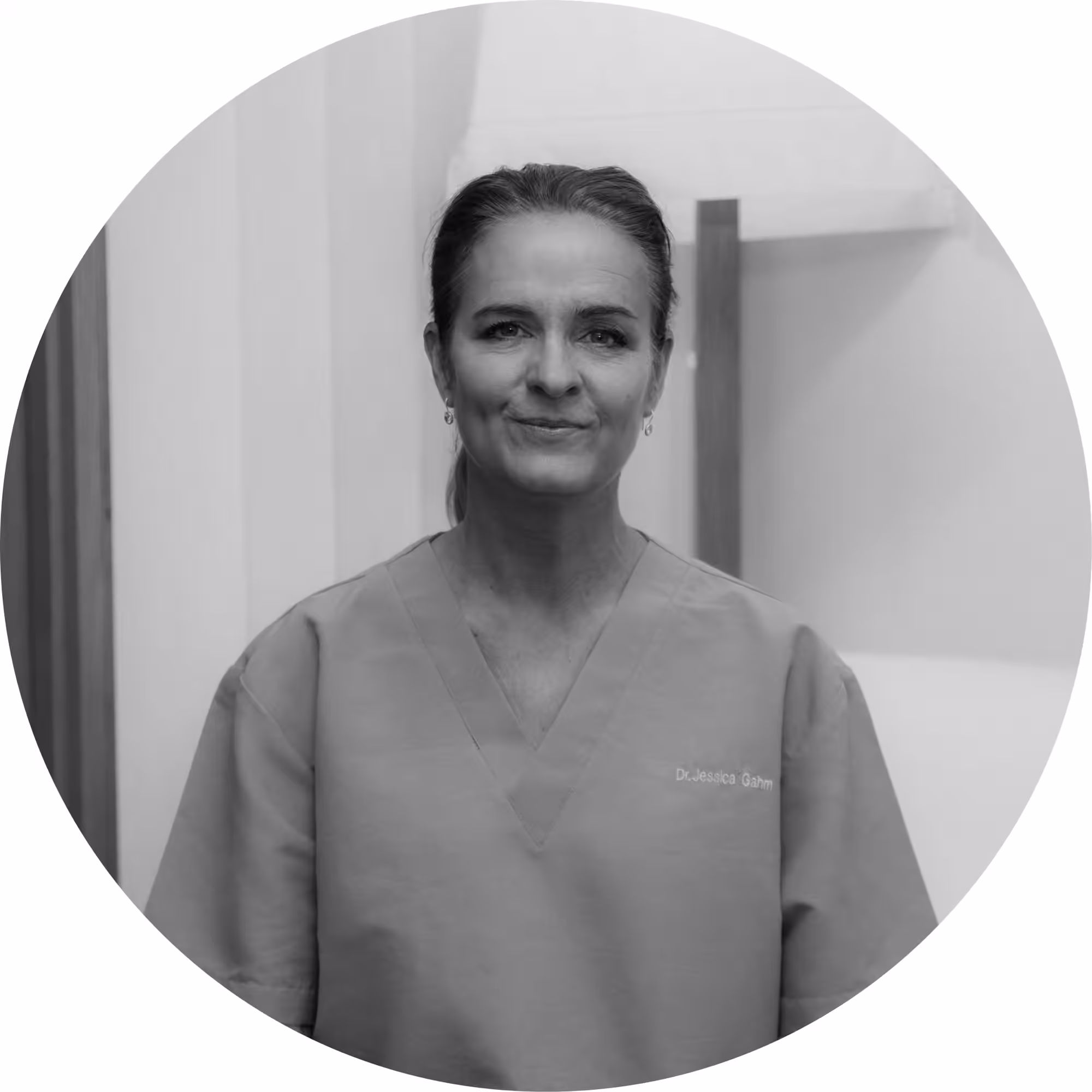 Black and white circular portrait photo of Dr. Jessica Garm, a woman wearing medical scrubs with her name embroidered on the chest, hair pulled back, smiling gently, photographed in a clinical hallway setting.