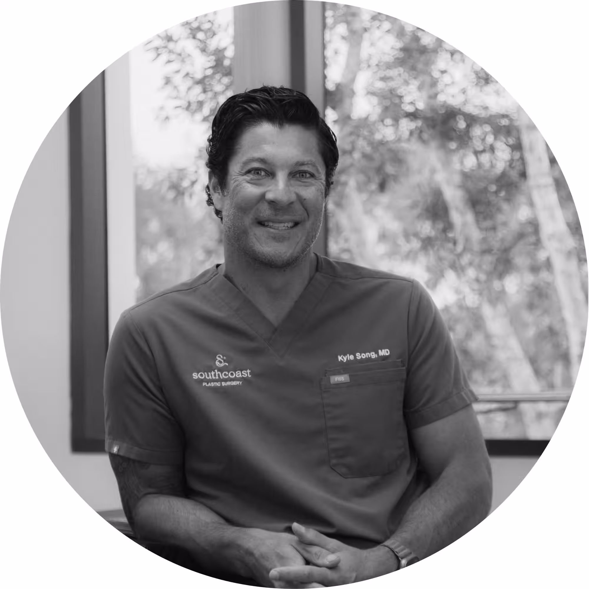 Black and white circular portrait photo of Dr. Kyle Song, MD, a smiling male plastic surgeon wearing medical scrubs embroidered with the South Coast Plastic Surgery logo and his name, seated with hands clasped, with a blurred window and trees visible in