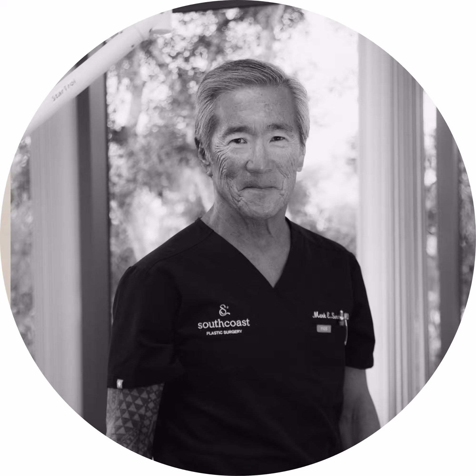 Black and white circular portrait photo of an older Asian man wearing dark medical scrubs embroidered with 'Southcoast Plastic Surgery' and a name tag reading 'Mark E. Su...', standing near a window with blurred trees in the background.