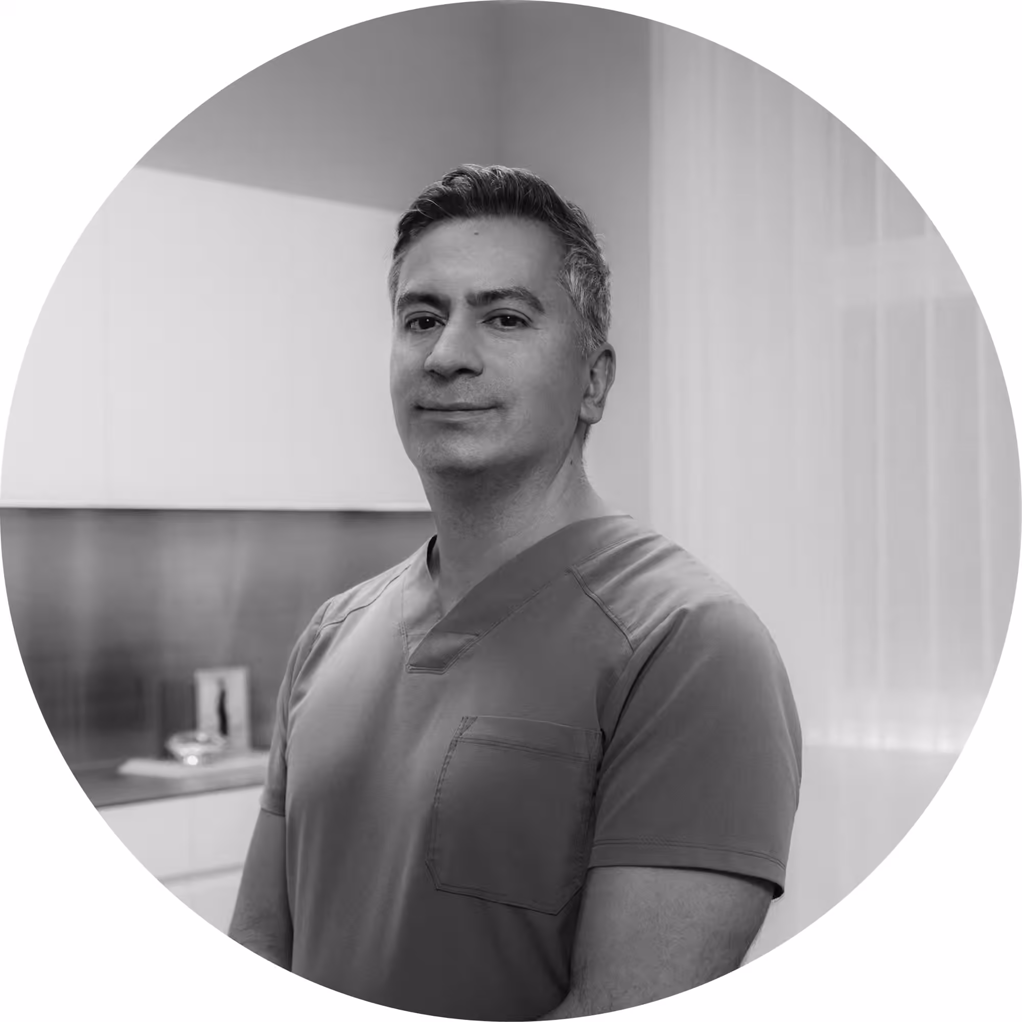 Black and white circular portrait photo of a middle-aged man with short salt-and-pepper hair, wearing a V-neck medical scrub top, standing in a clean clinical or medical office setting.