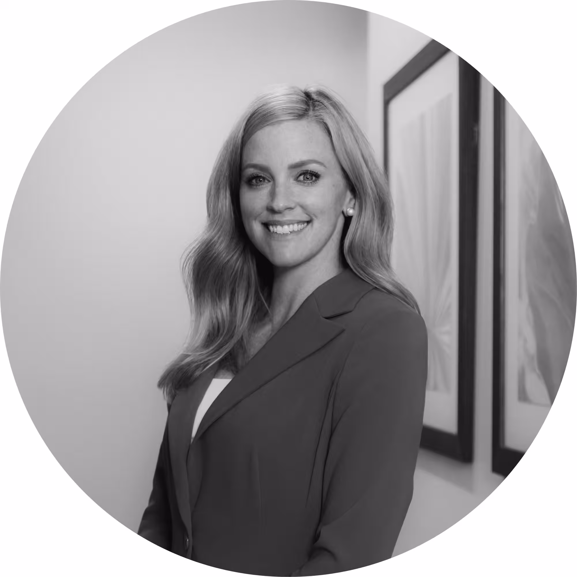 Black and white circular professional headshot of a smiling woman with long wavy blonde hair, wearing a blazer, posed in front of a wall with framed artwork.