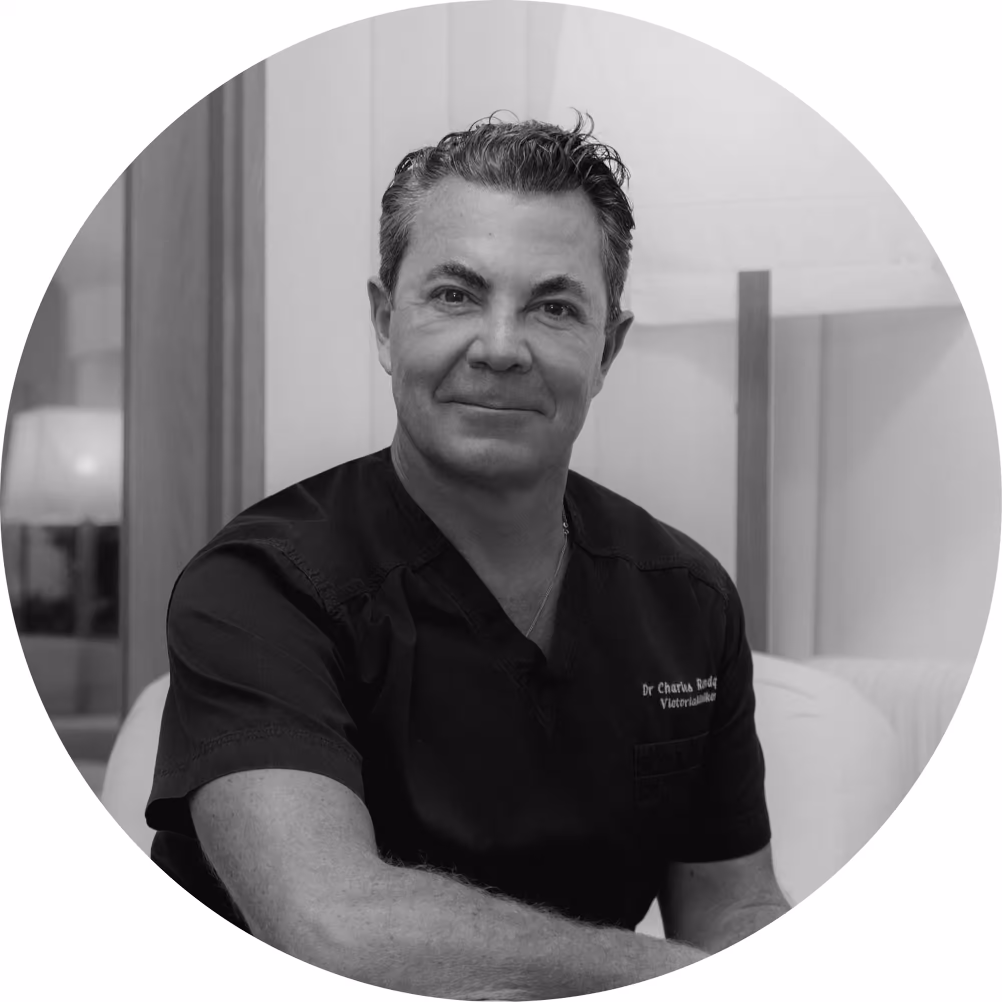 Black and white circular portrait photo of Dr. Charles Rody, a male medical professional with dark hair wearing dark scrubs with his name embroidered on the chest, seated and smiling in a clinical setting.