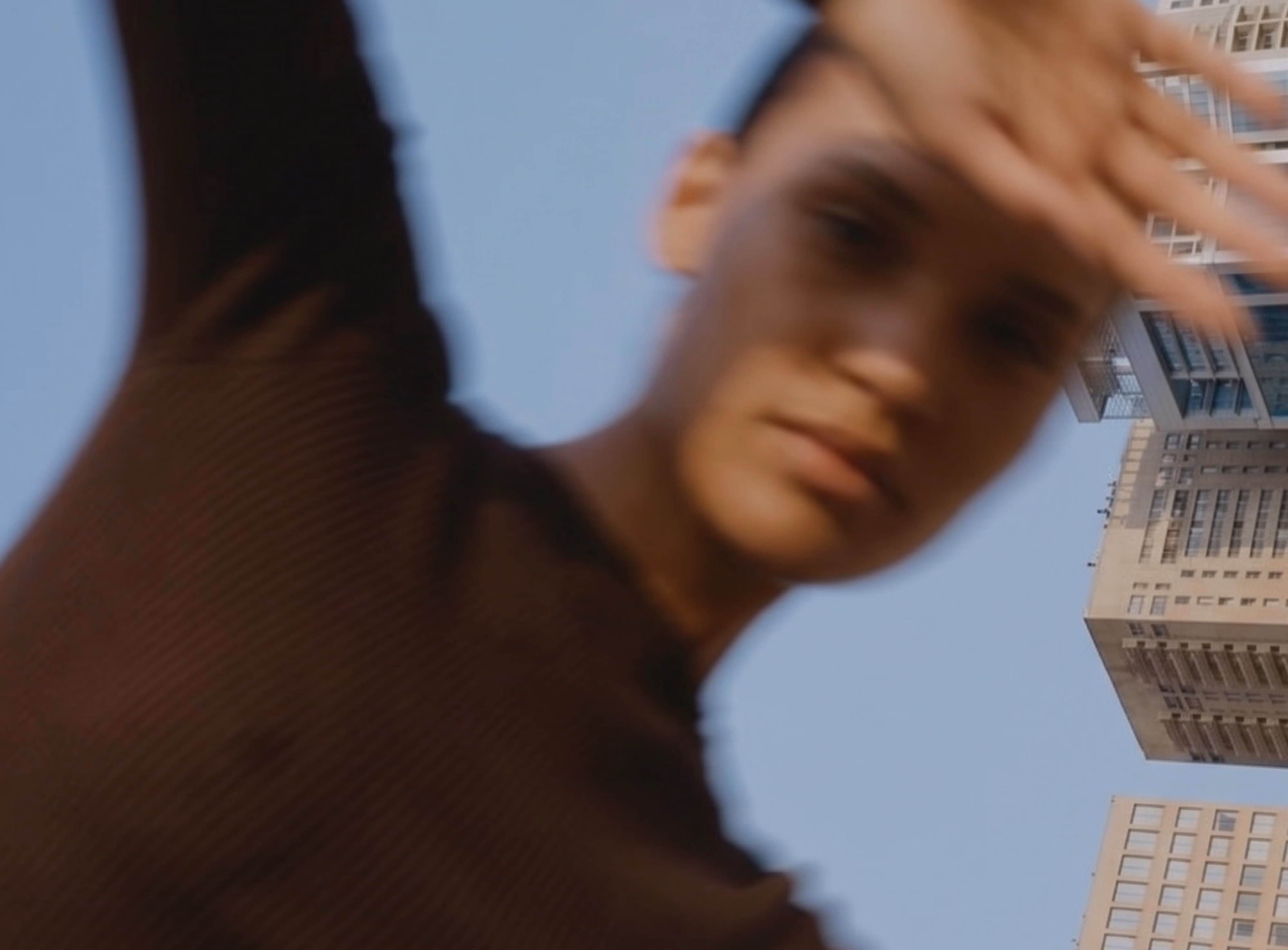 A motion-blurred artistic photo of a young man in a dark brown long-sleeve top, with one arm raised and a hand in motion near his head, shot from a low angle against a clear blue sky with modern high-rise buildings visible in the background.