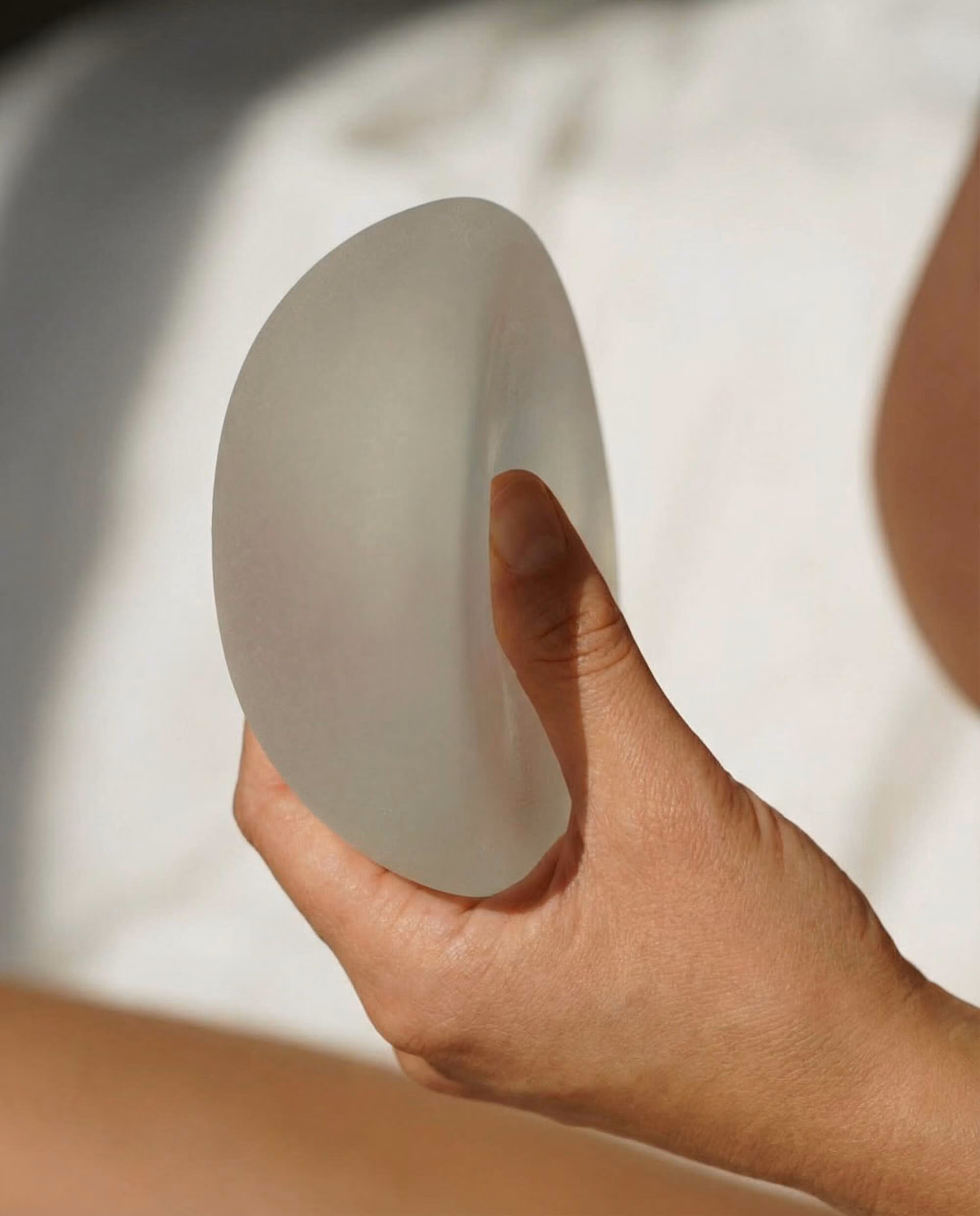 A hand holding a translucent silicone breast implant against a white fabric background, with sunlight casting soft shadows.