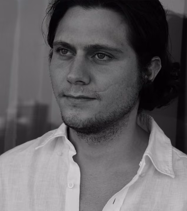Black and white close-up portrait of a young man with dark shoulder-length hair, light stubble, and brown eyes, wearing an open-collar white linen shirt, with a softly blurred background.