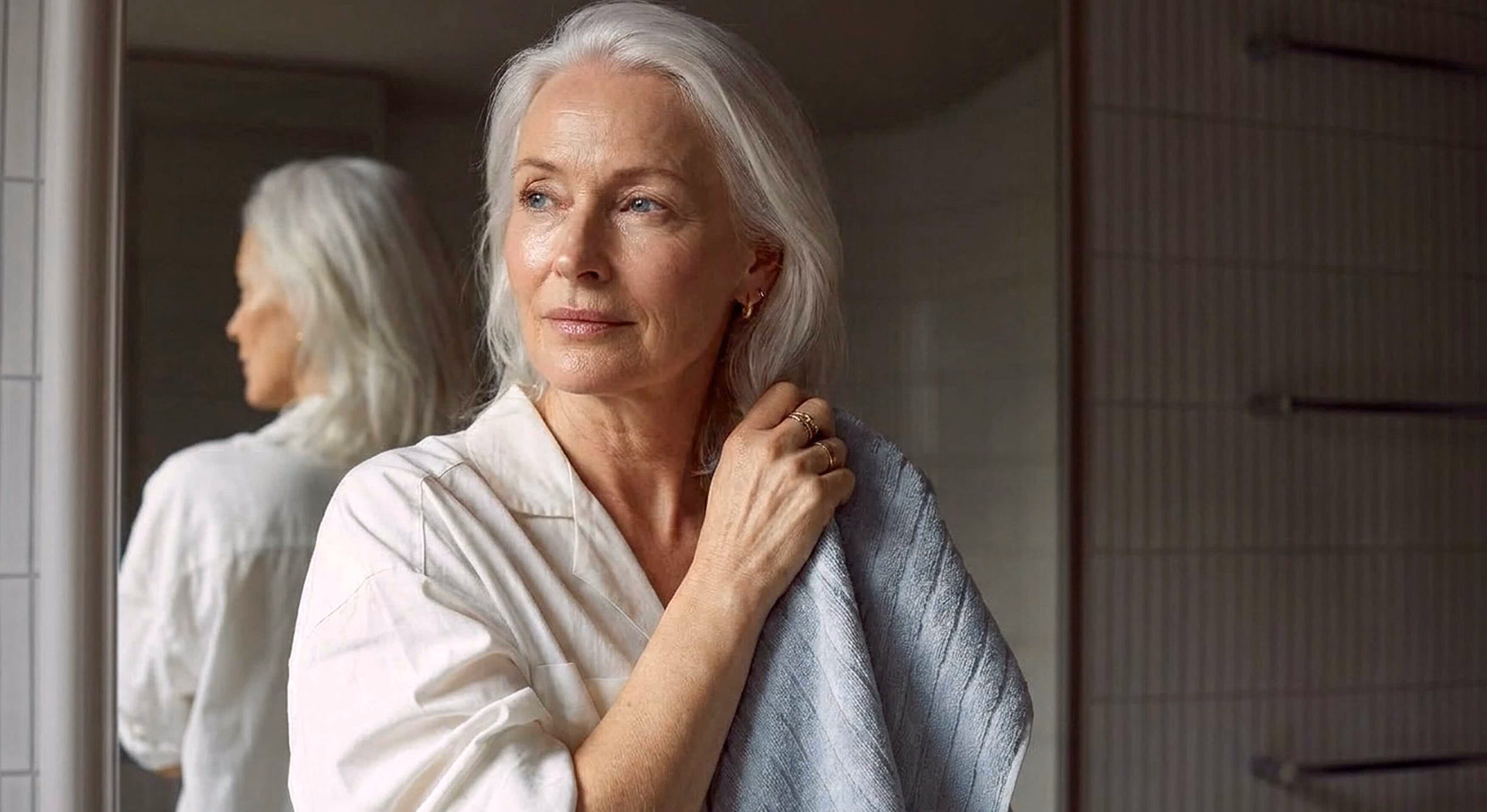 An older woman with long silver-gray hair wearing a white robe, holding a gray towel to her neck in a bathroom setting, with her reflection visible in a mirror behind her.