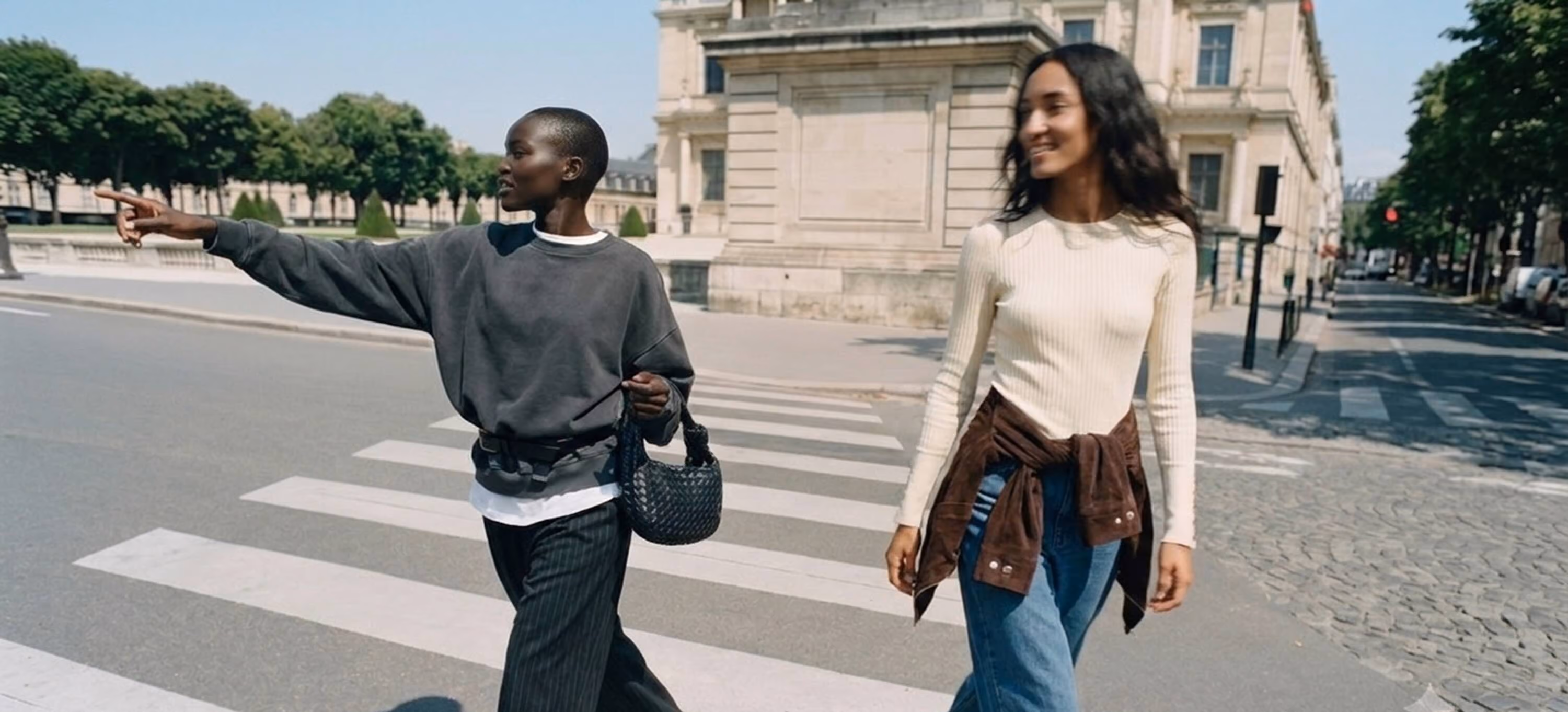 Two women walking confidently across a pedestrian crosswalk on a sunny Parisian street. The first woman wears a gray oversized sweatshirt, wide-leg dark trousers, and carries a small woven black handbag, with one arm outstretched pointing ahead. The