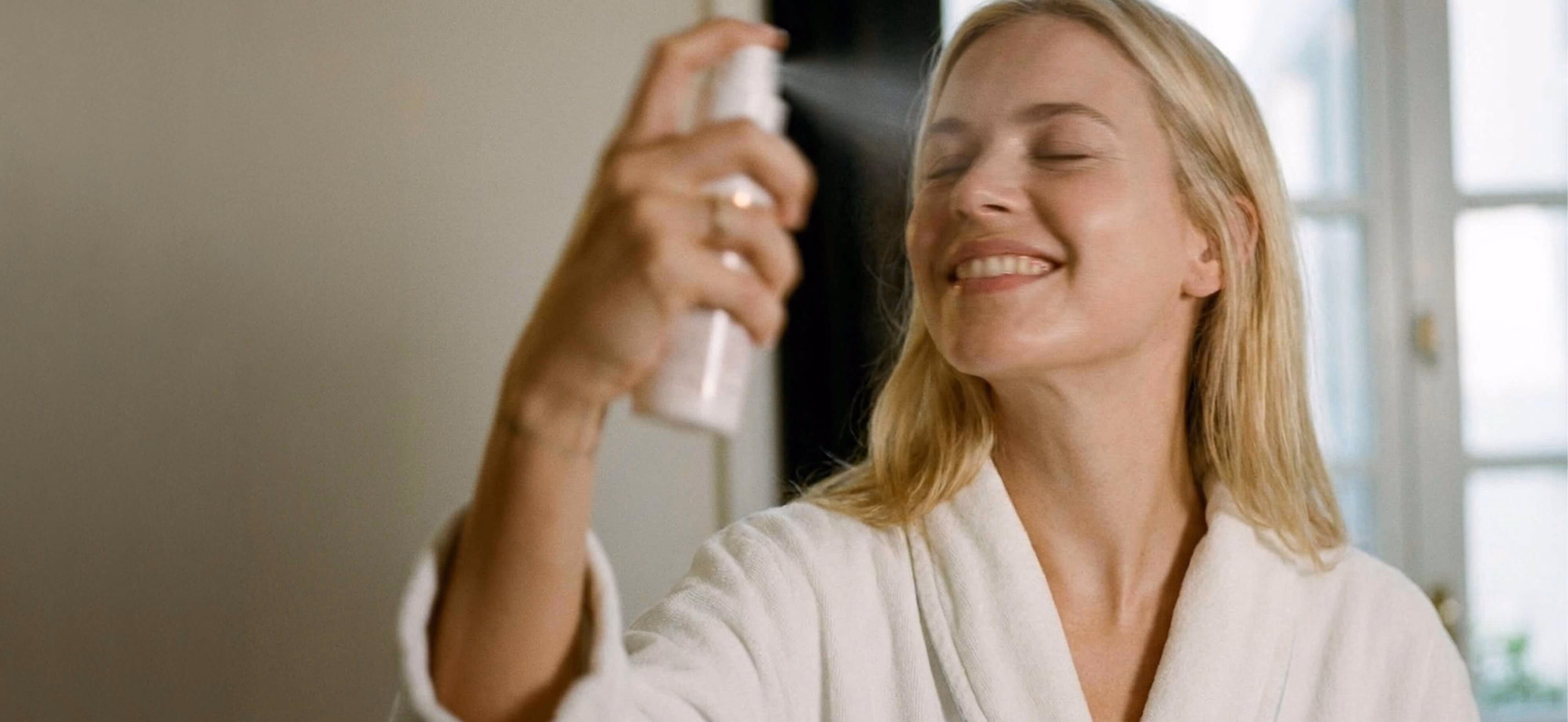 A smiling woman with blonde hair, wearing a white bathrobe, spritzing a facial mist spray from a light pink aerosol bottle toward her face with her eyes closed.