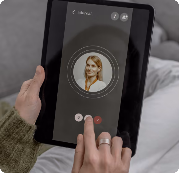 A person holding a tablet displaying the Adoreal app interface, showing a smiling woman's circular profile photo with concentric ring animations, a microphone button, and a red close button, suggesting an active AI voice or video call session.