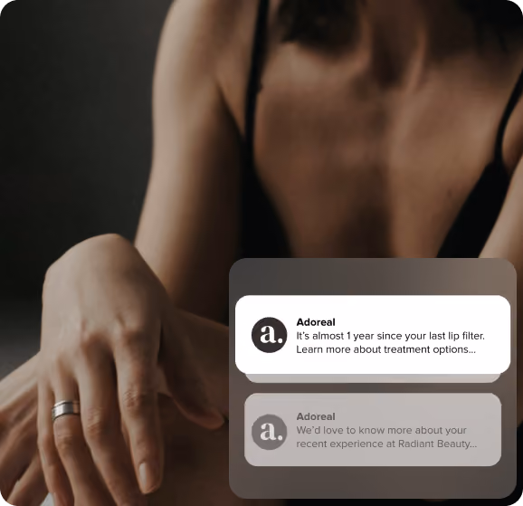 A woman resting her arms forward wearing a black top and a silver ring, with two overlaid app notification cards from Adoreal. The top card reads: 'It's almost 1 year since your last lip filter. Learn more about treatment options…' and the second card
