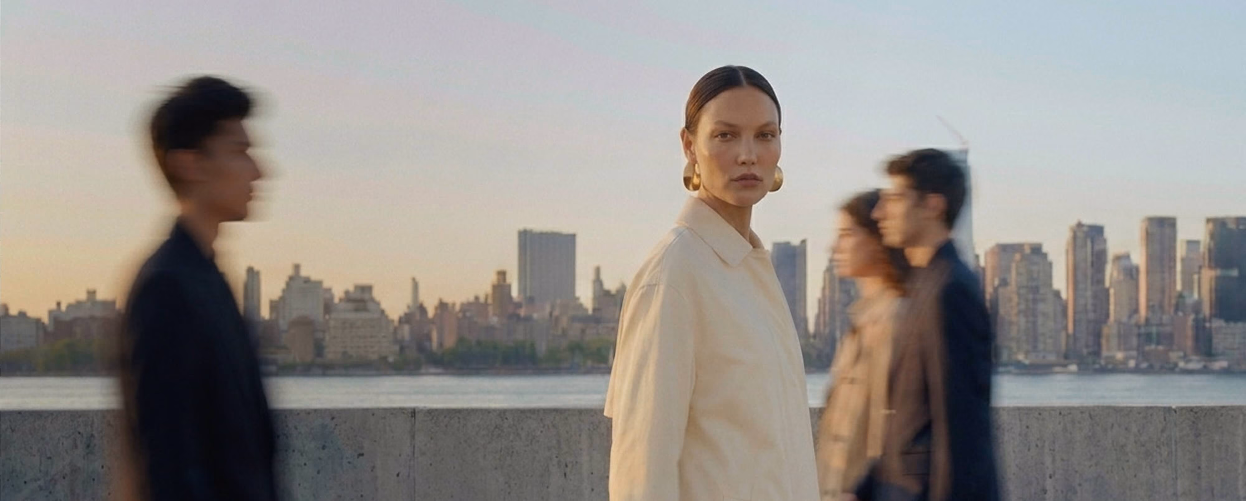 A sharp-focused woman in a cream oversized coat stands still on a waterfront promenade, with motion-blurred pedestrians passing around her, and a city skyline across the river under a soft dusk sky in the background.