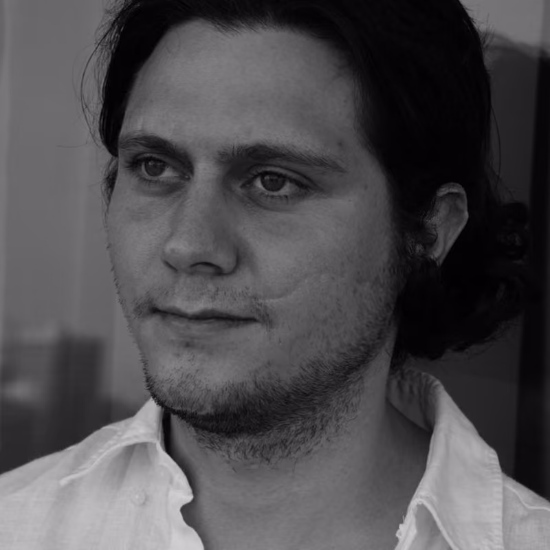 Black and white portrait photo of a young man with shoulder-length dark hair tied back, light stubble beard, and wearing an open-collar white shirt, gazing slightly off-camera.