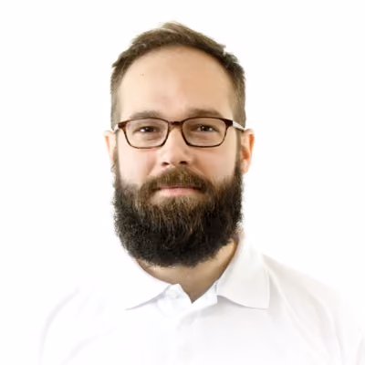 Professional headshot of a man with a full dark beard and mustache, wearing rectangular-framed glasses and a white collared shirt, smiling slightly toward the camera.