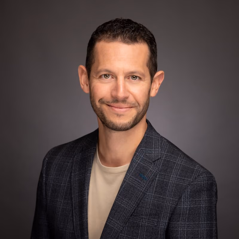Professional headshot of a man with short dark hair and light stubble, smiling warmly at the camera. He is wearing a dark navy plaid blazer over a beige crew-neck t-shirt.