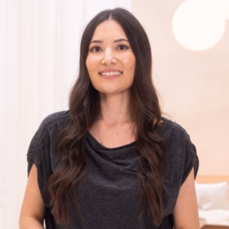 A smiling woman with long, wavy dark brown hair wearing a dark gray short-sleeve top, standing in a bright, softly lit interior space with a round pendant light visible in the background.