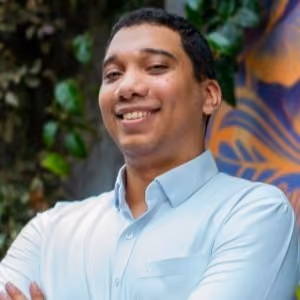 A smiling young man with short hair, wearing a light blue button-up shirt and crossing his arms, with green foliage and a colorful mural visible in the background.